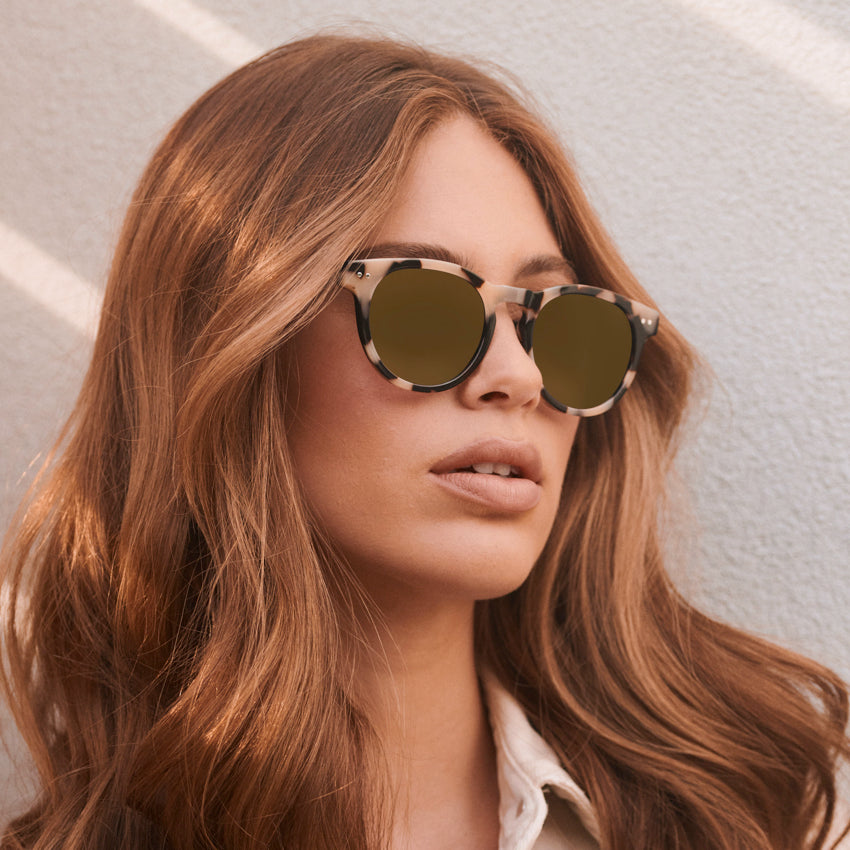 Woman wearing retro round-shaped sunglasses with tortoiseshell frame and double rivet details against a light background
