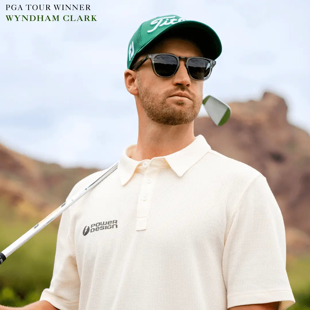 Man wearing performance sunglasses and green golf cap holding golf club on course with rocky background