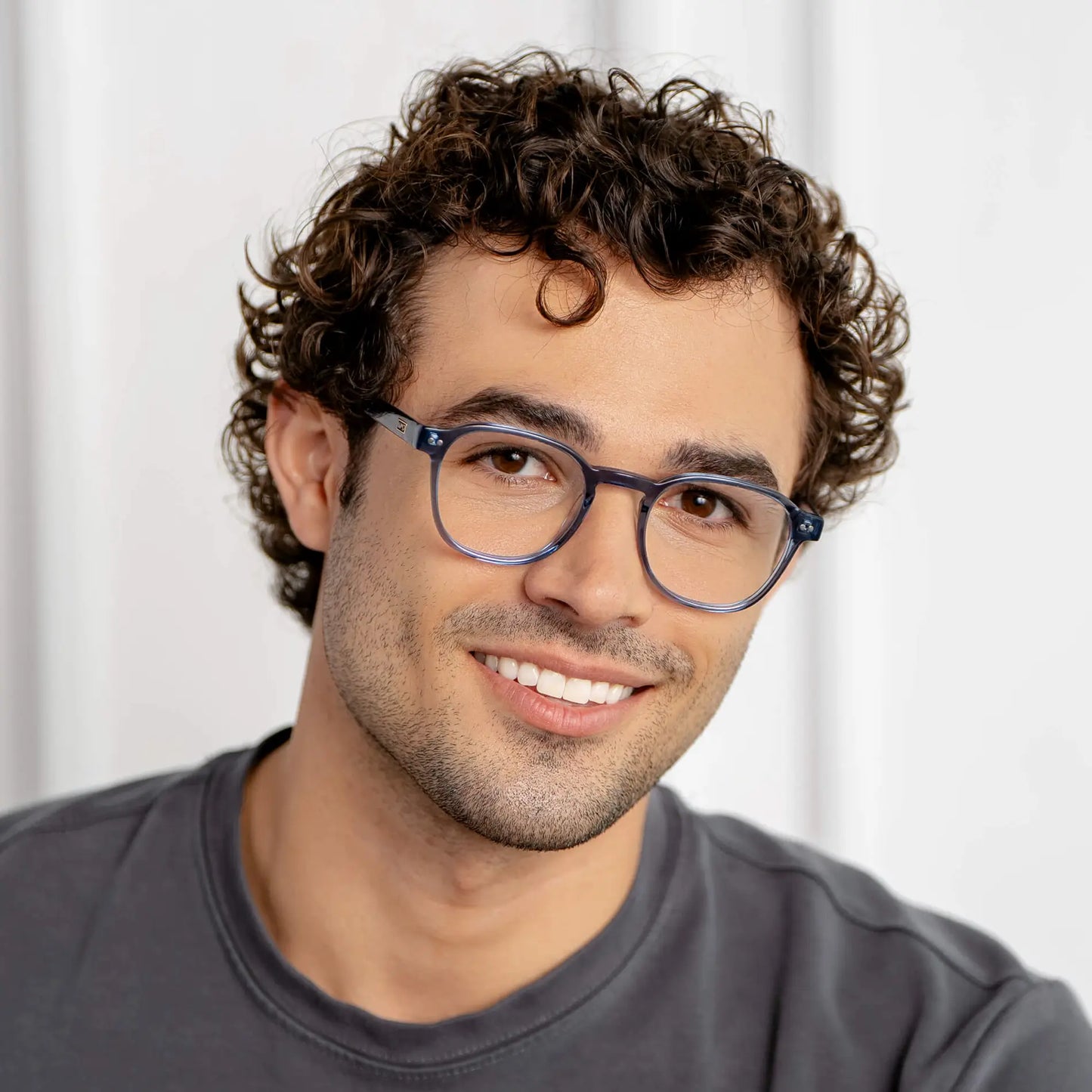 Pacific Blue || Chase blue light glasses with transparent blue rounded frames worn by smiling man with curly hair in natural lighting