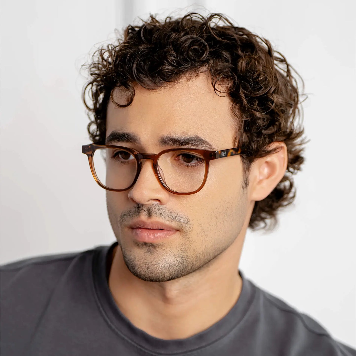 Crystal Brown || Side view of rounded square prescription eyeglasses with crystal brown frame and clear lens worn by man with curly hair indoors