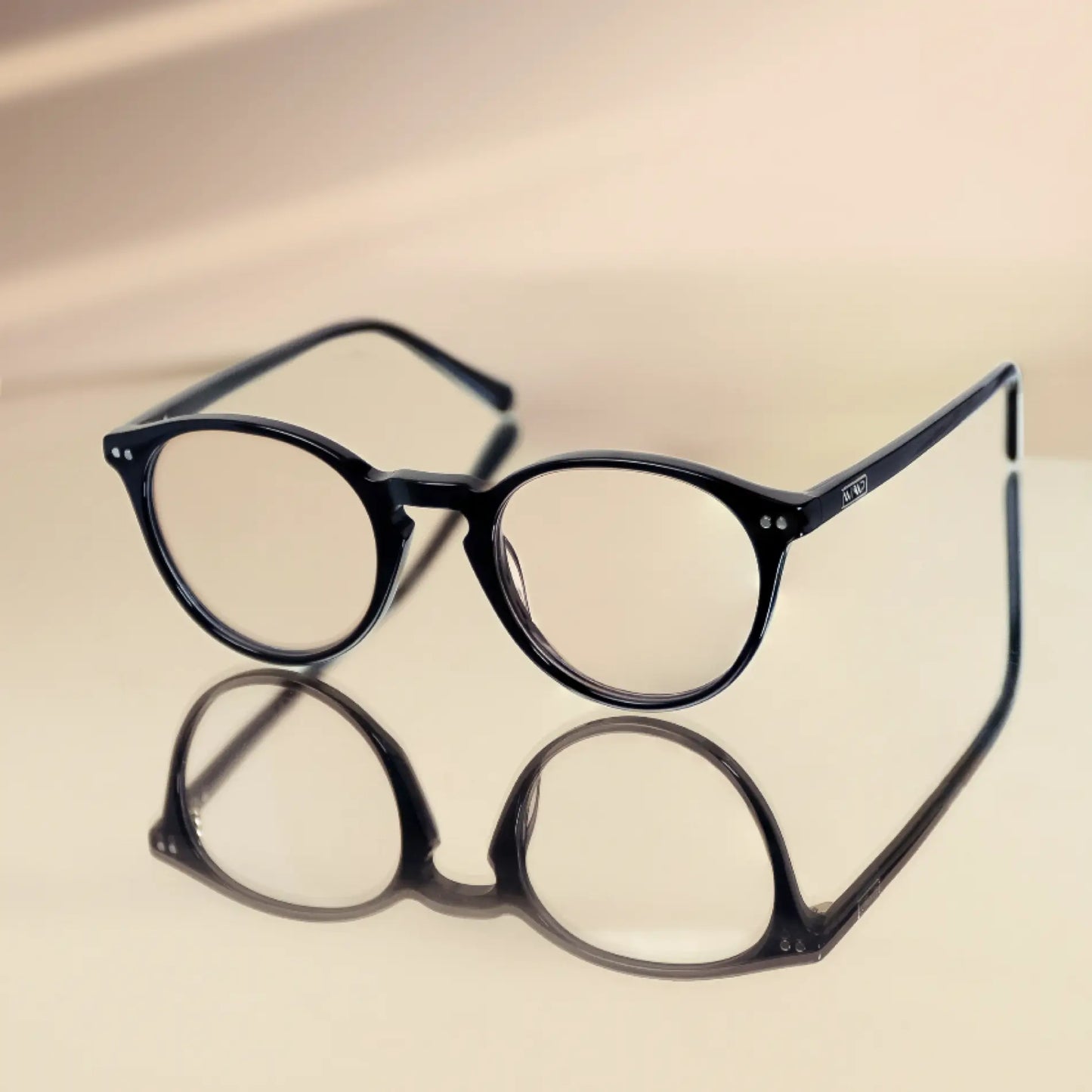 Glossy Black || Black rounded frames blue light glasses resting on reflective surface with soft neutral background