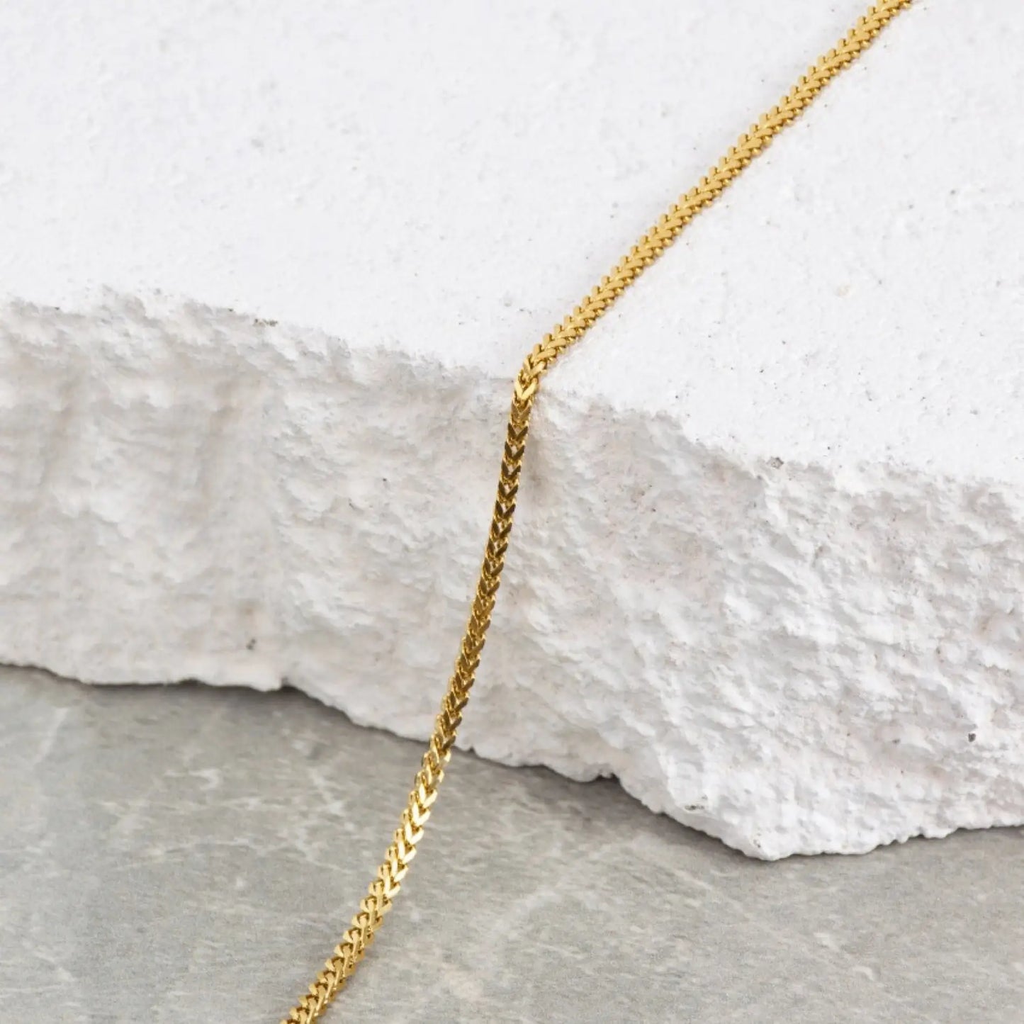 Gold || Gold tone stylish chain draped over rough white stone surface with gray background