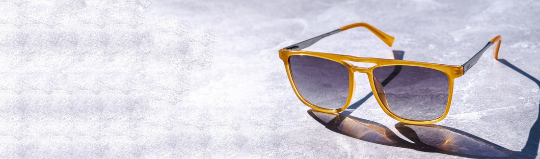 Single pair of double bridge aviator sunglasses with translucent amber acetate frames and gradient gray lenses resting on a bright textured stone surface in natural sunlight