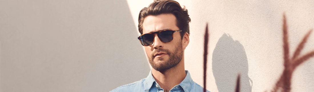 Man wearing classic black wayfarer sunglasses with dark lenses, a light blue denim shirt, styled dark hair and beard, posed against a warm beige wall with soft plant shadows