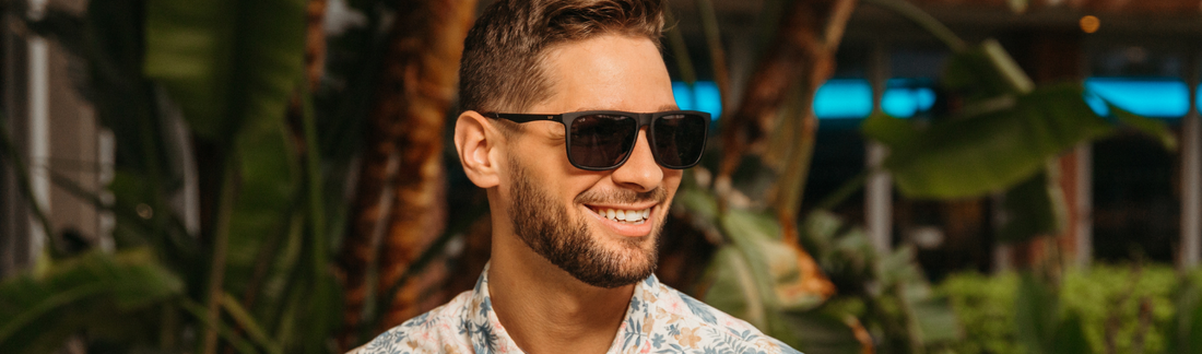 Smiling man wearing square tortoiseshell aviator-style sunglasses with dark lenses, a floral print shirt, with short hair and beard, photographed outdoors among tropical palm trees