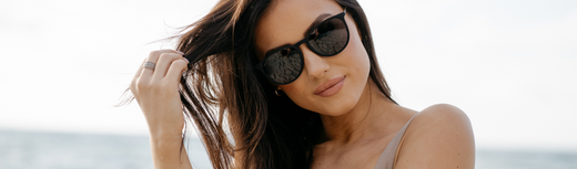 Woman wearing black sunglasses by the ocean, touching her hair in natural light