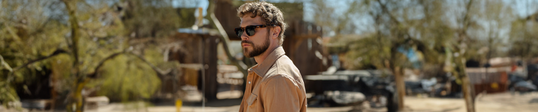 Man with short curly hair and beard wearing black rectangular sunglasses and a tan shirt, looking over his shoulder outdoors in a sunny desert setting