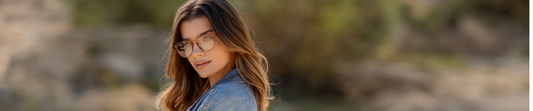 Woman with long brown hair wearing brown frame glasses, looking over her shoulder outdoors with a blurred natural background