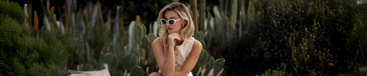 Woman wearing sunglasses, seated outdoors with cactus plants in the background