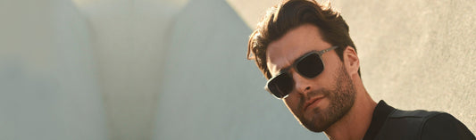 Man wearing square light grey frame sunglasses with black lenses, dressed in a black shirt, with styled dark hair and beard, photographed against a soft blue-gray outdoor wall