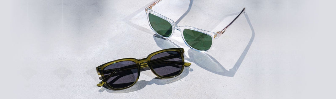 Two pairs of sunglasses on a light stone surface: a oversized cat-eye style with clear crystal frames and green lenses, and a square frame with olive green acetate and dark gray lenses, casting natural shadows in a flat lay