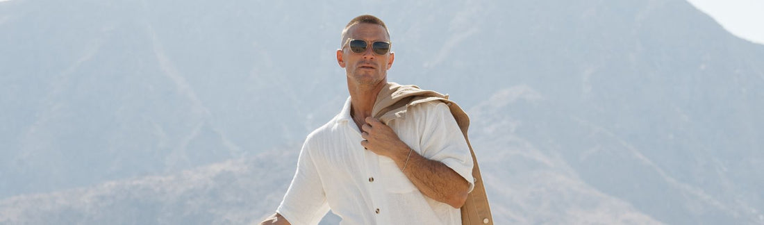 Man wearing sunglasses outdoors holding a jacket over his shoulder with mountains in the background