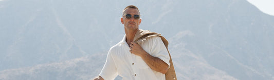 Man wearing sunglasses outdoors holding a jacket over his shoulder with mountains in the background