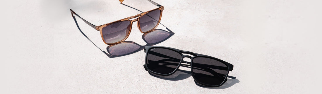 Two square aviator style sunglasses on light concrete: amber frames with gradient gray lenses, and matte black frames with solid black lenses, in natural sunlight