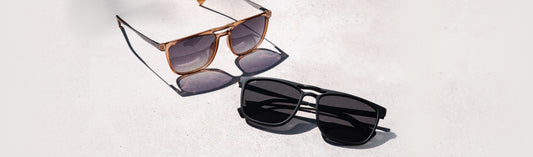 Two square aviator style sunglasses on light concrete: amber frames with gradient gray lenses, and matte black frames with solid black lenses, in natural sunlight