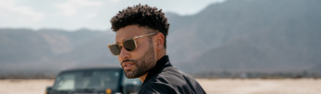 Man wearing a black jacket and square yellow frame sunglasses outdoors with a mountains in the background