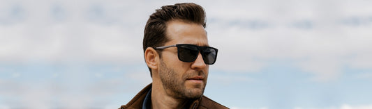 Man wearing classic black wayfarer sunglasses with dark lenses, a brown leather jacket, and blue shirt, with styled dark hair and beard, photographed against a cloudy sky