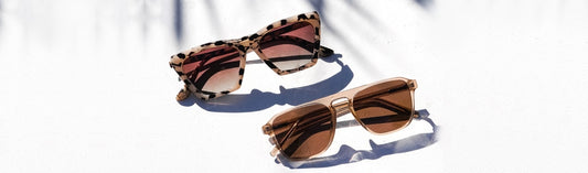 Two pairs of sunglasses with brown lenses on a bright white surface with palm shadow overlay: a cat-eye style with black and white tortoiseshell acetate frames, and a square browline style with warm tan acetate and metal frames, shot in natural sunlight