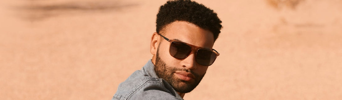 Man wearing square tortoiseshell aviator-style sunglasses with brown lenses, dressed in a denim jacket, glancing back over his shoulder against a warm sandy desert background