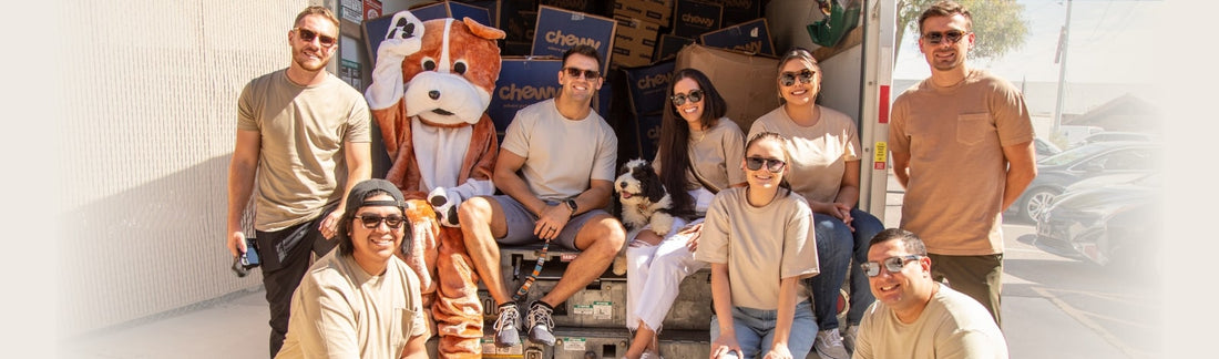 Group of seven people wearing sunglasses posing in front of a delivery truck alongside the dog mascot and a live dog, in a sunny outdoor brand event setting
