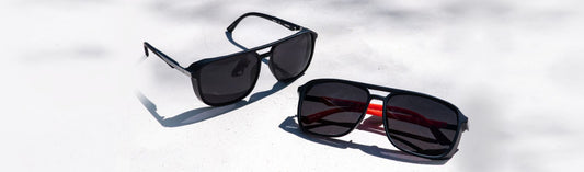 Two pairs of sunglasses with black aviator frames resting on a light surface in natural light.