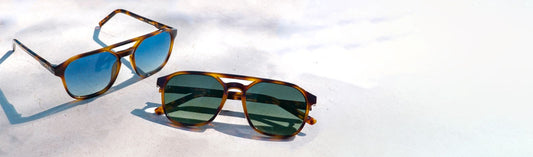 Two pairs of sunglasses with green and blue lenses displayed on a sunlit surface in natural light