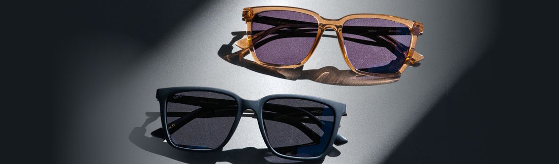 Two pairs of square acetate sunglasses on a dark surface featuring amber frames with purple-tinted lenses, and matte navy blue frames with gray lenses, casting shadows in an editorial flat lay