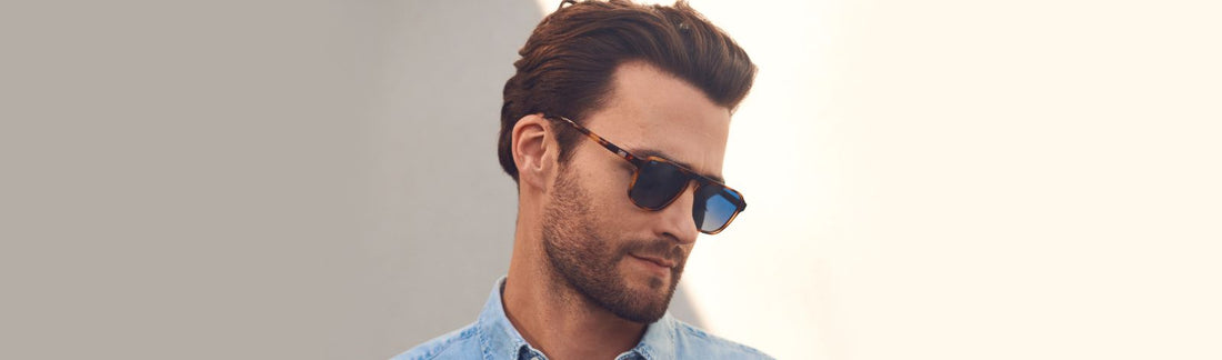 Man wearing aviator browline sunglasses with tortoiseshell frames and blue gradient lenses, dressed in a light blue denim shirt with short styled brown hair and beard, looking downward against a bright outdoor background