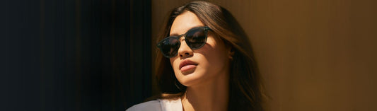 Woman wearing classic black sunglasses with dark lenses and a light gray top, with straight dark hair, gazing upward in dramatic directional sunlight against a warm-toned wall in a fashion lifestyle setting