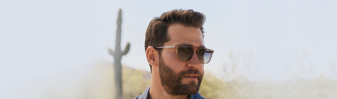 Man wearing rectangular frame sunglasses with gradient brown lenses, dressed in a blue shirt with styled brown hair and beard, photographed outdoors against a bright desert sky with a saguaro cactus in the background