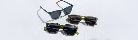 Three pairs of square acetate sunglasses with dark gray lenses arranged on a white linen surface: one translucent emerald green frame and two olive green frames in varying sizes, casting natural shadows in a flat lay product shot