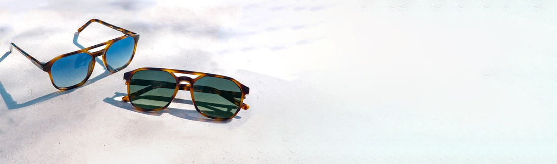 Two pairs of sunglasses with blue and green lenses displayed on a sunlit surface in natural light.