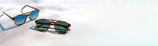 Two pairs of sunglasses with blue and green lenses displayed on a sunlit surface in natural light.