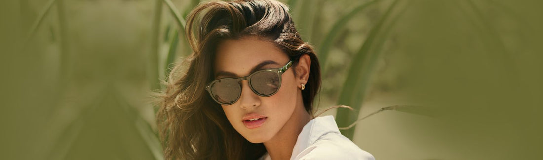 Woman wearing round tortoiseshell sunglasses with brown lenses and a white open-collar shirt, with long wavy dark hair, posed against a lush green tropical plant background in a fashion lifestyle setting