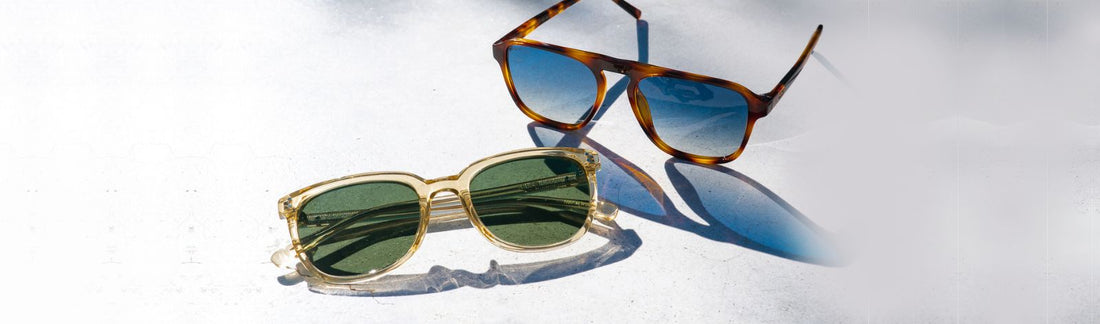 Two pairs of sunglasses with blue and green lens resting on a light surface in natural light.