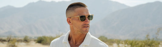 Man wearing sunglasses with green lens outdoors with mountains in the background.