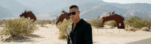 Man wearing sunglasses walking outdoors in a desert landscape with metal sculptures in the background