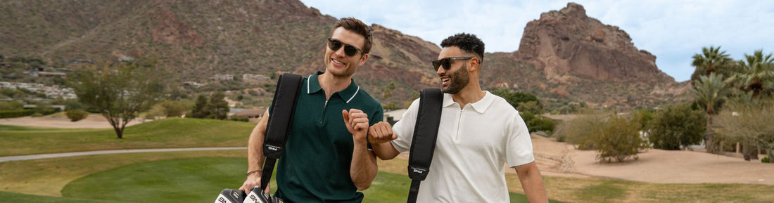 Two men wearing sunglasses walking outdoors with golf bags, desert mountains in the background