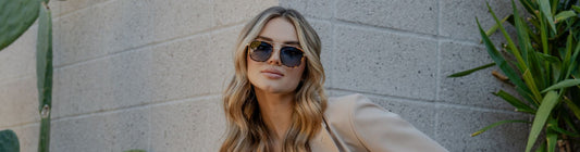 Woman wearing oversized aviator sunglasses standing against a textured wall outdoors.