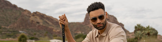Man wearing sunglasses holding a golf club on a course with desert mountains behind him