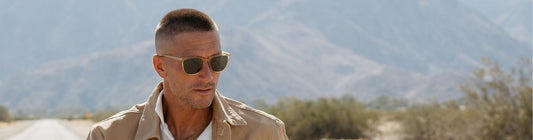 Man wearing sunglasses outdoors with plants and mountains in the background.