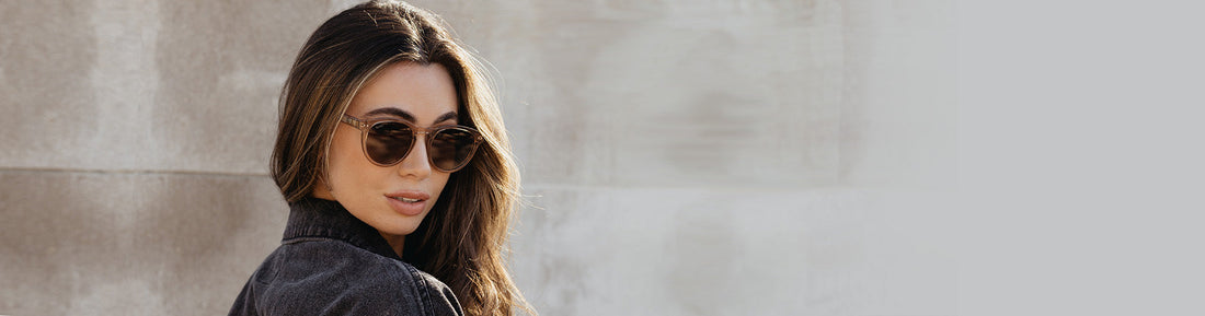 Woman wearing sunglasses looking over her shoulder against a neutral wall.
