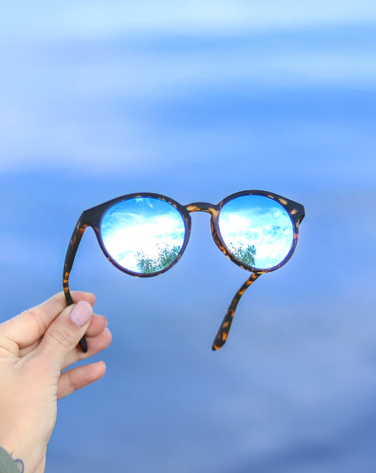Hand holding up round tortoiseshell sunglasses with blue mirrored lenses reflecting trees and sky, held against a bright blue cloudy sky background
