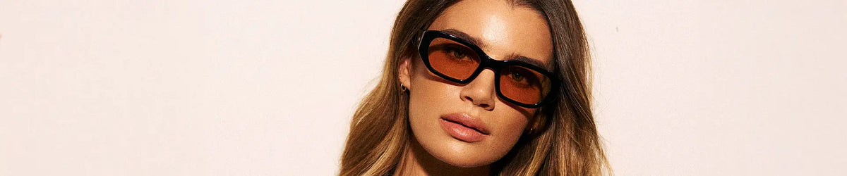 Woman wearing black rectangular WMP London style sunglasses with gold-tinted lenses, cream background.