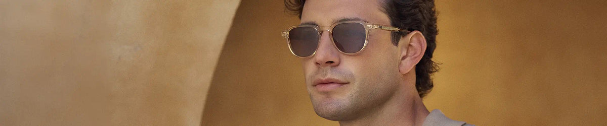 Man wearing translucent sand brown round sunglasses against a warm tan background.