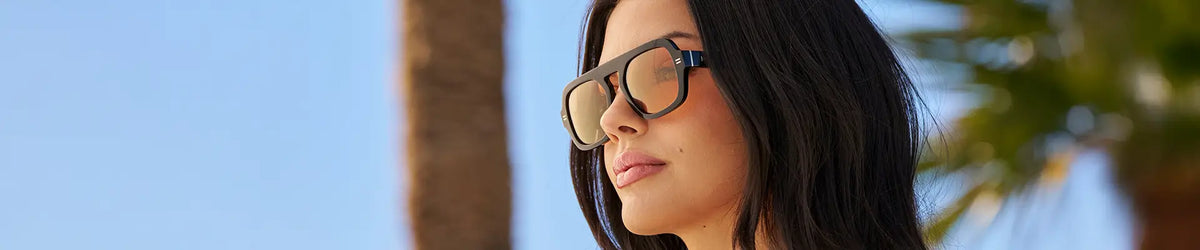Woman wearing black aviator-frame WMP Naomi sunglasses with orange lens, profile view, palm trees and blue sky background. 