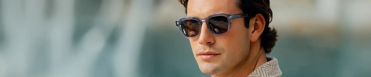 Man wearing demim blue geometric-frame WMP Nelson sunglasses, brown lenses, blurred outdoor background.