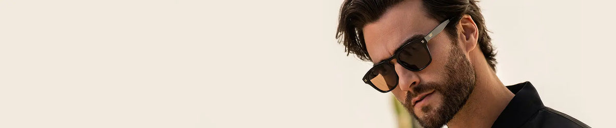 Close-up of a bearded man wearing dark rectangular sunglasses and a black shirt, looking downward against a light, neutral background.