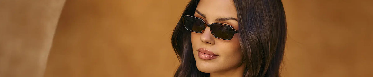 Woman wearing slim brown tortoise rectangular sunglasses against a warm tan background.