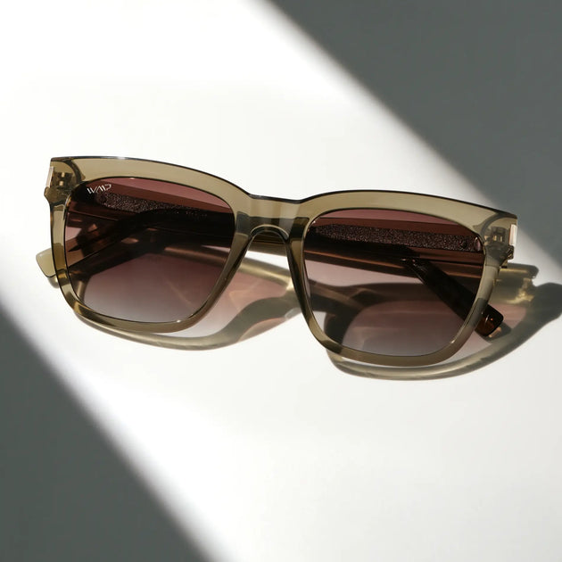 Folded pair of square sunglasses with translucent olive-green frames and dark brown lenses resting on a white surface in directional sunlight.